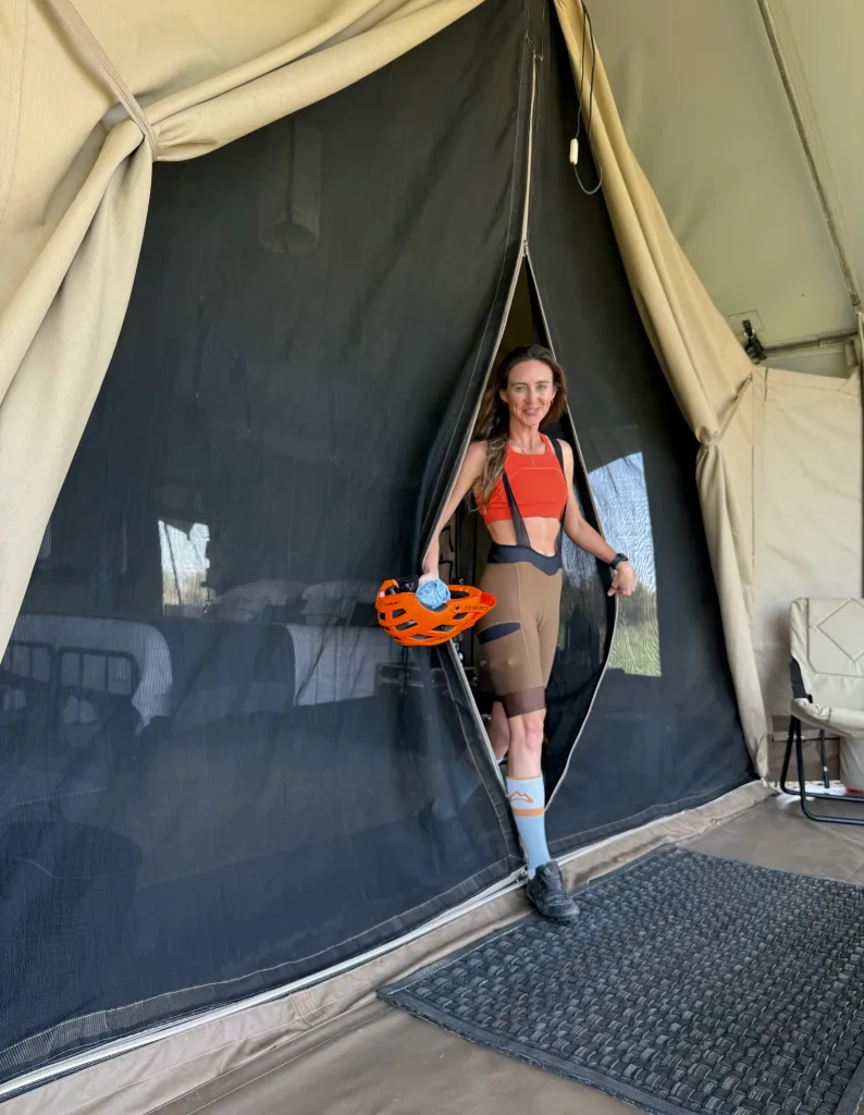 A woman in cycling gear exits a safari tent with an orange helmet.