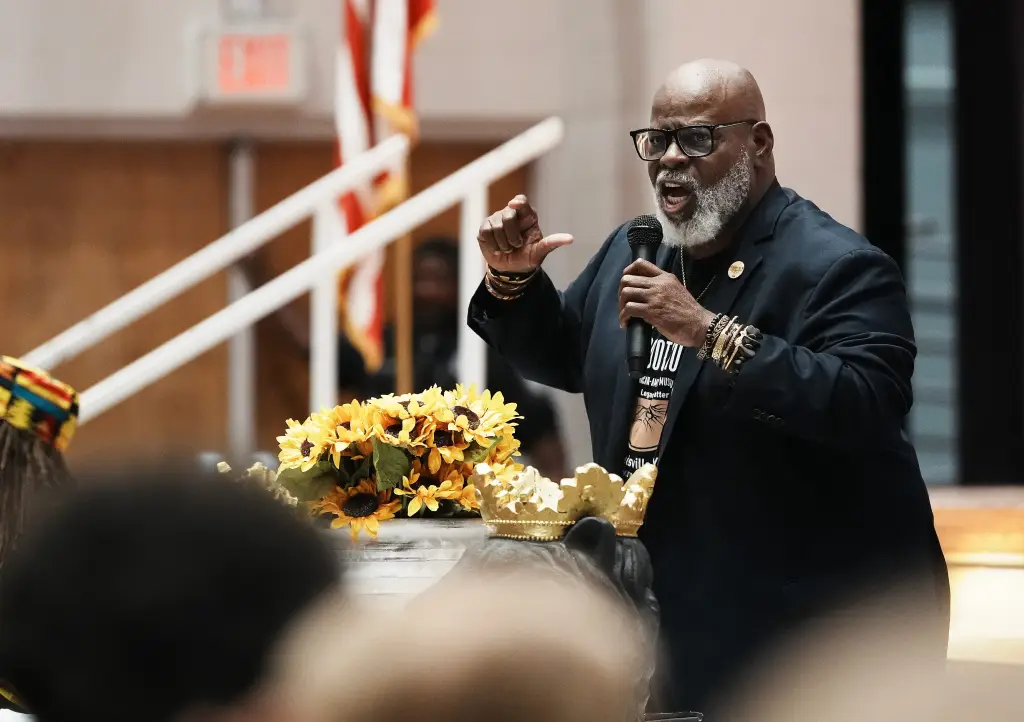 A man with a white beard and glasses speaks into a microphone, gesturing with his hand.