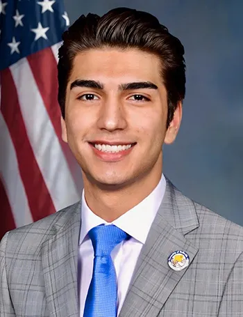 Headshot of Velez in a suit and tie, smiling in front of an American flag.