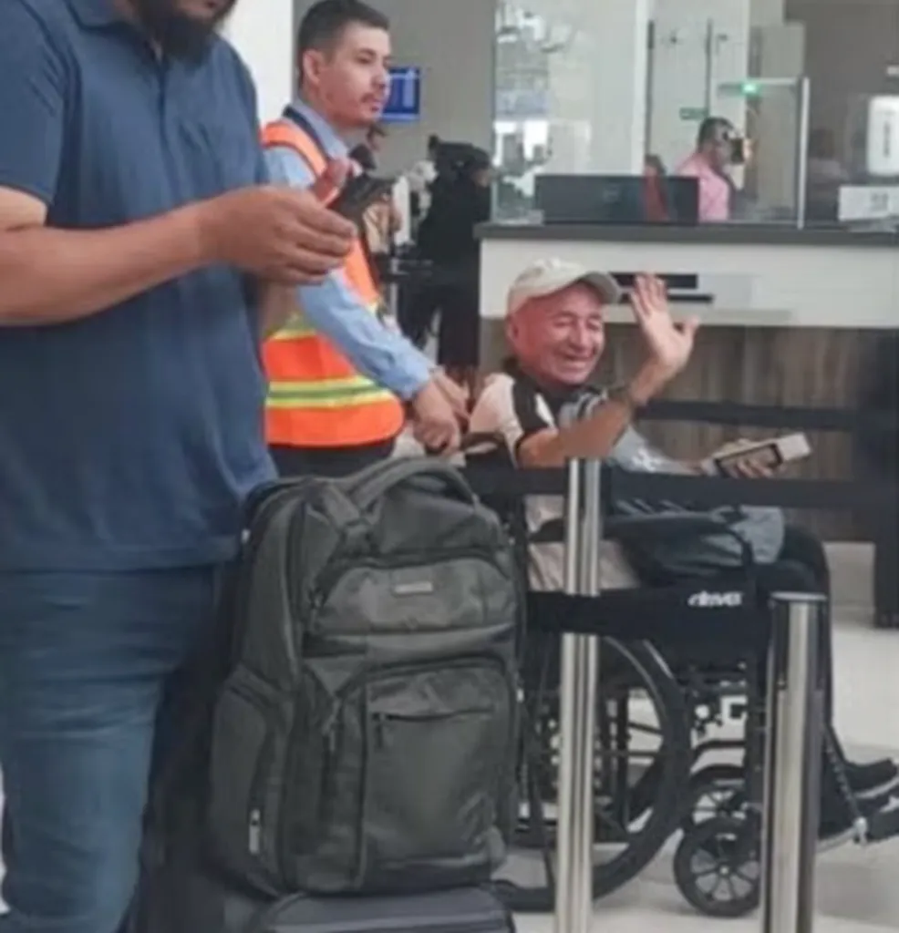 A man in a wheelchair waves, accompanied by airport personnel and another man, next to a backpack.