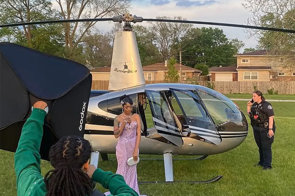 A woman in a prom dress posing in front of a helicopter for photos, with a photographer holding a softbox and a police officer nearby.