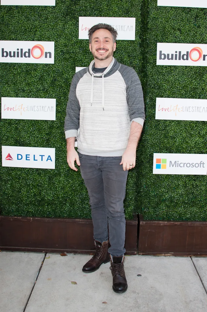 Actor Adam Mondschein smiles while posing in a white and gray hoodie, dark jeans, and brown boots in front of a green backdrop with various logos.