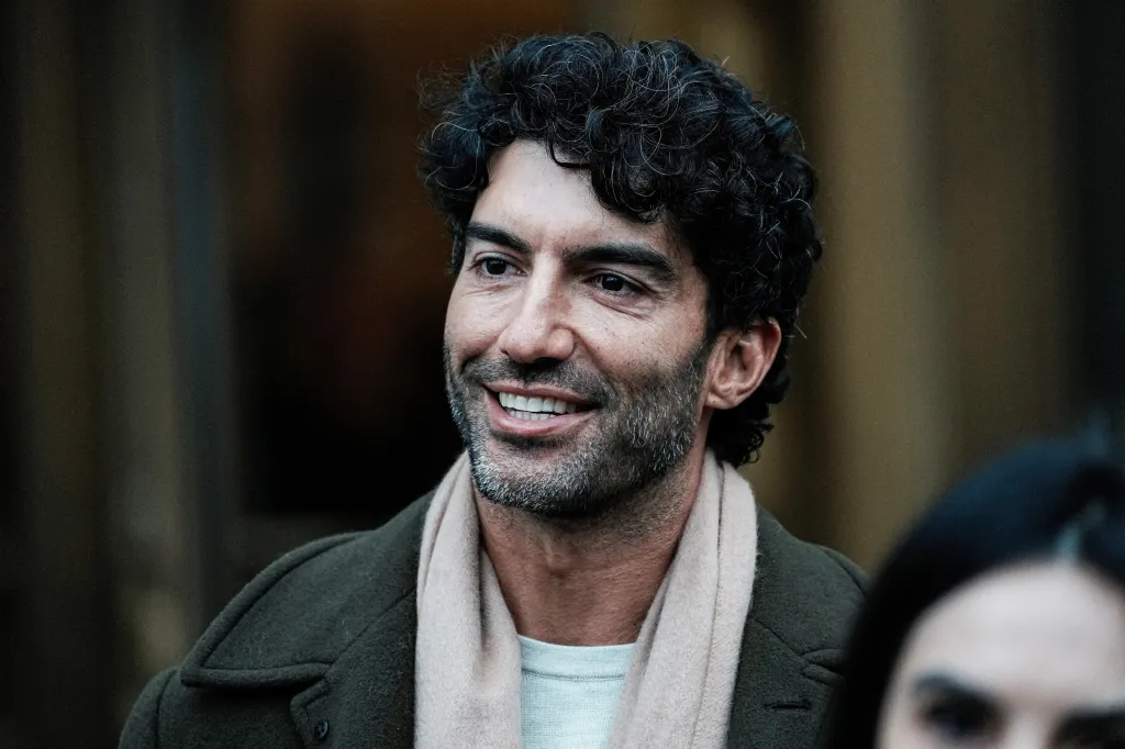 Actor Justin Baldoni smiles, dressed in a dark green coat and light pink scarf.