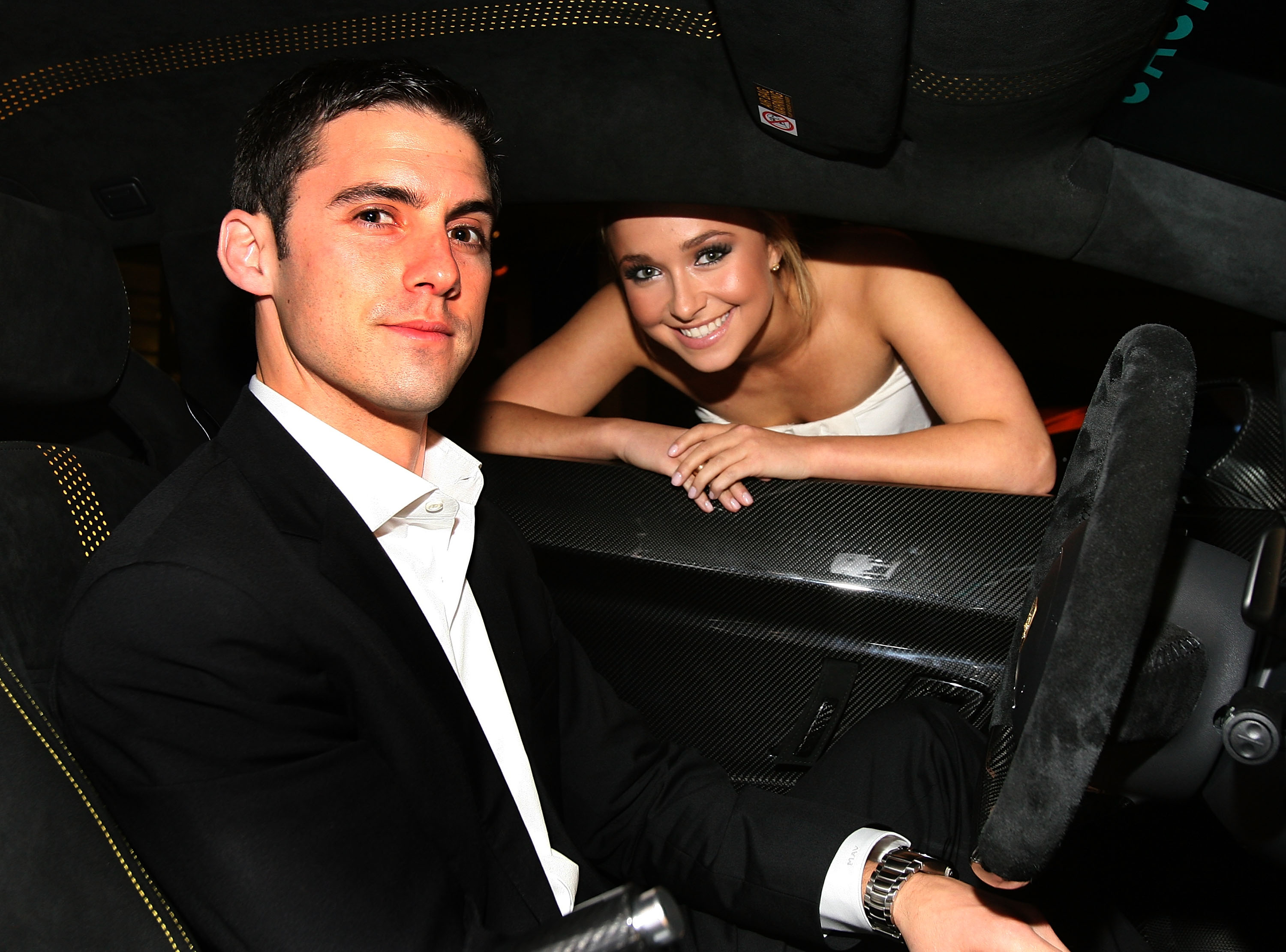 Milo Ventimiglia in the driver's seat of a car, looking at the camera, with Hayden Panettiere leaning into the car from the passenger side, smiling at the camera.