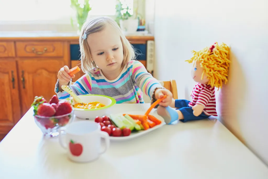 Toddler girl eating fresh fruits and vegetables while feeding her doll.