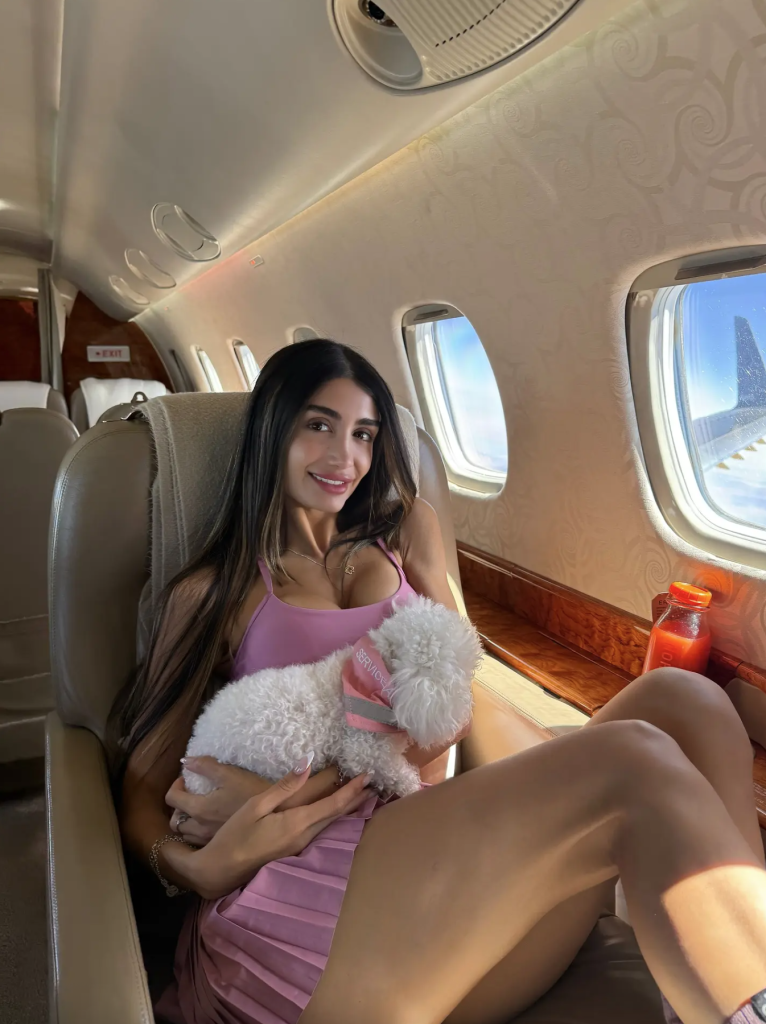 Adva Lavie, also known as Mia Ventura, seated in a private jet holding a small white dog with a pink