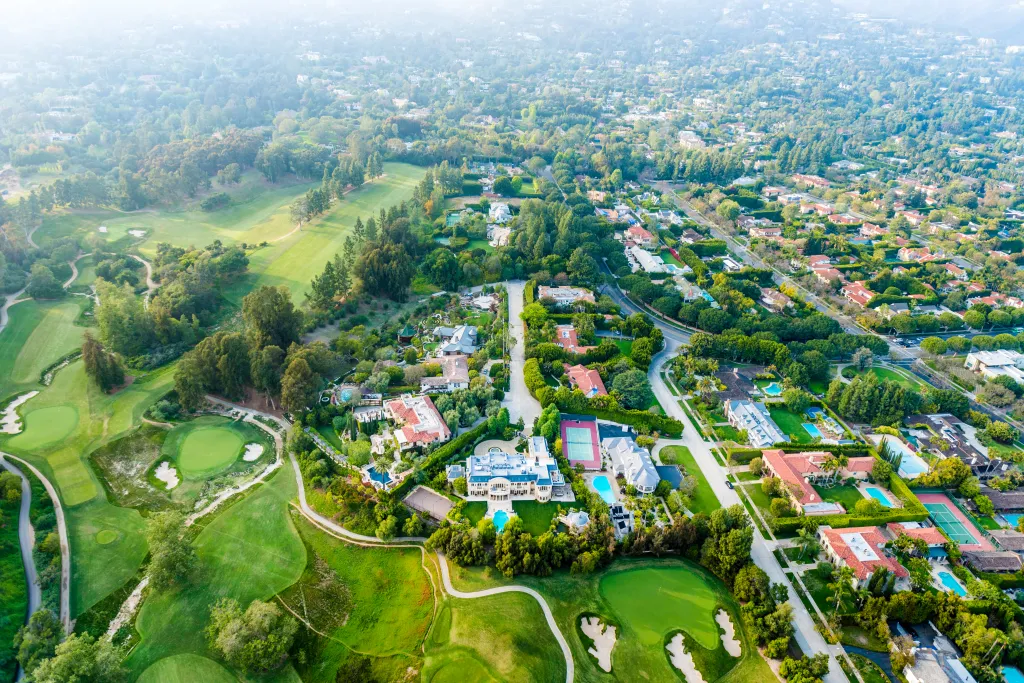 Aerial view of the Bel Air neighborhood in Los Angeles, showing mansions, roads, and a golf course under hazy skies.