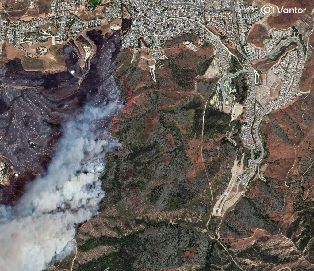 Aerial satellite view of damage from the Sandy Fire, showing burned land and smoke next to a residential area.