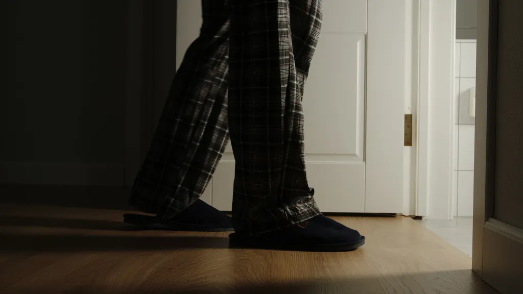 An aged man in plaid pajamas and slippers walks to a toilet at home in the night.