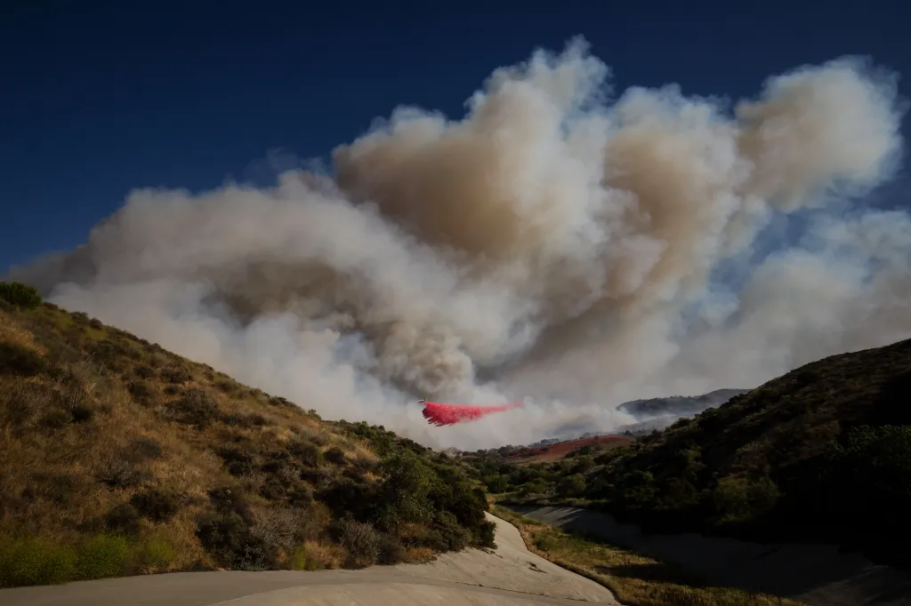 An air tanker drops red fire retardant on a wildfire in a hilly, brush-covered landscape, producing a large plume of smoke.