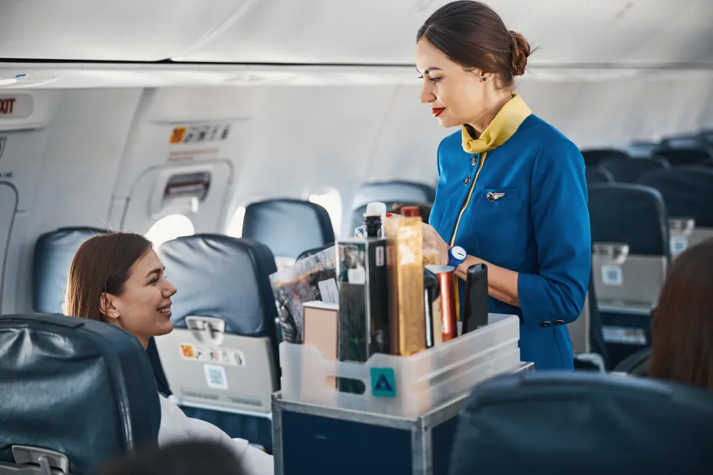 A flight attendant in a blue and yellow uniform speaks to a female passenger from behind a food trolley in an airplane aisle.