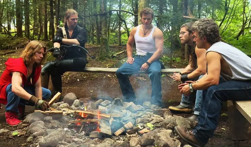 Five men gather around a campfire in the woods.