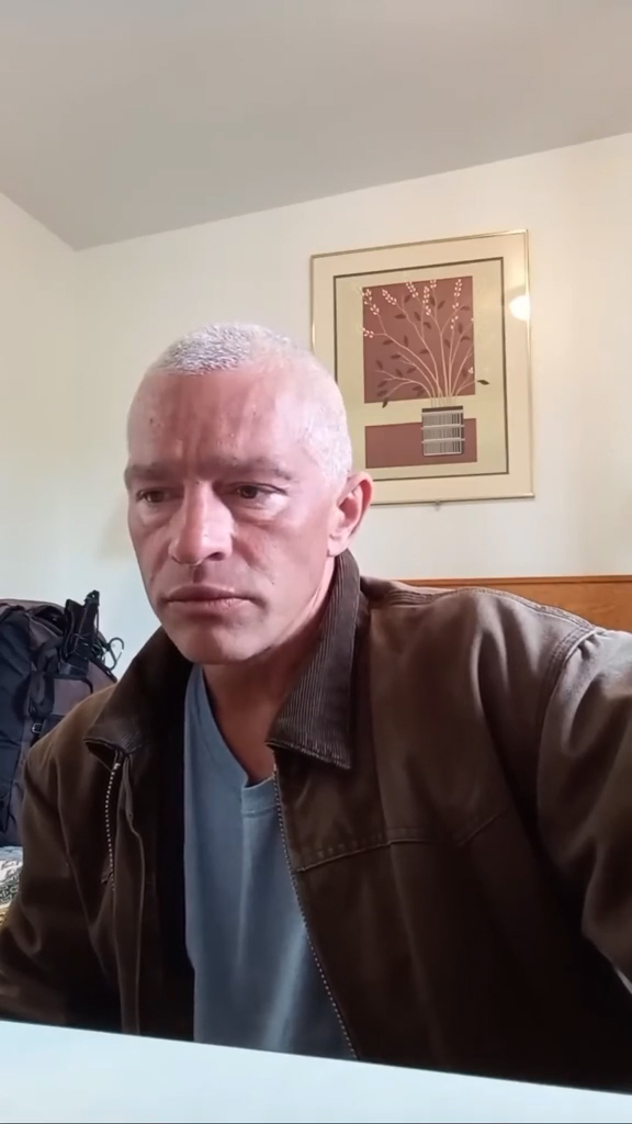 Matt Brown, with short white hair and a brown jacket, sits in a room with a framed picture on the wall.