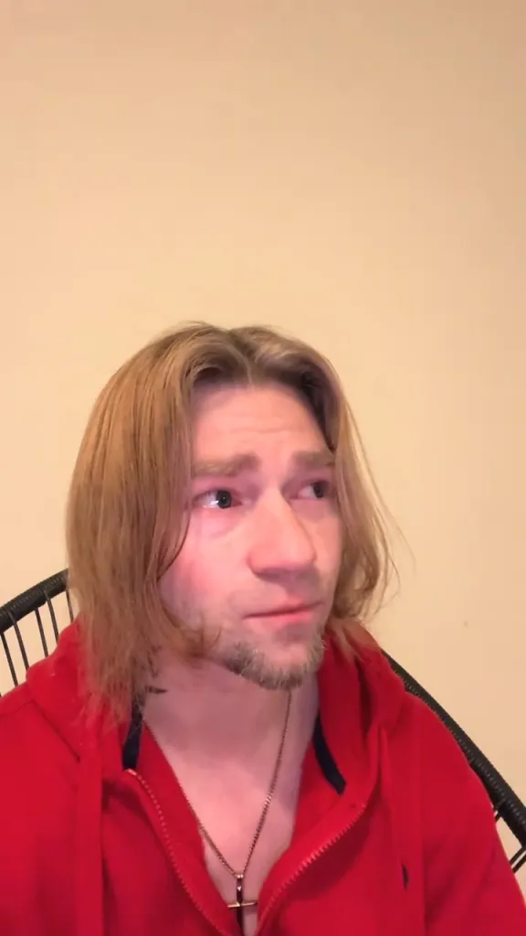 A close-up of a fair-skinned man with long, light brown hair, a goatee, and red-rimmed eyes, wearing a red hooded sweatshirt and a cross necklace.