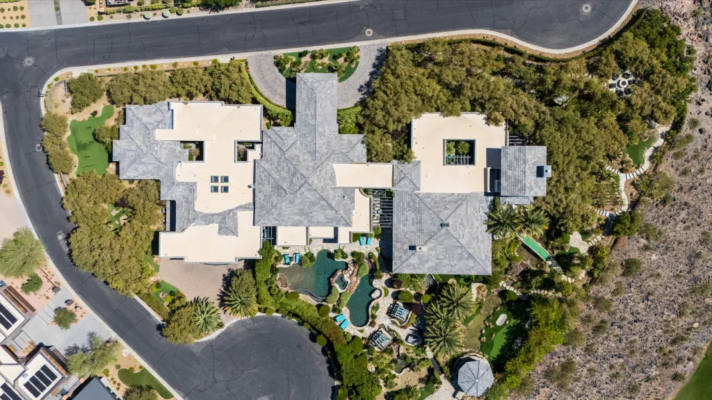 Aerial view of a sprawling luxury home with multiple buildings, swimming pools, a putting green, and lush landscaping, surrounded by roads and arid hills.