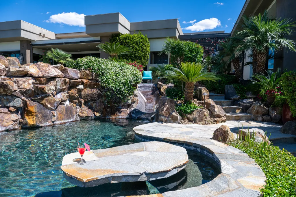 A luxury outdoor pool area with a waterfall, stone-paved walkway, and a red cocktail with a pink umbrella resting on a stone ledge.