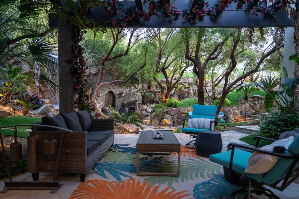 Outdoor patio with a sofa and two chairs, a colorful rug, and a lush backyard with a waterfall and a stone archway.