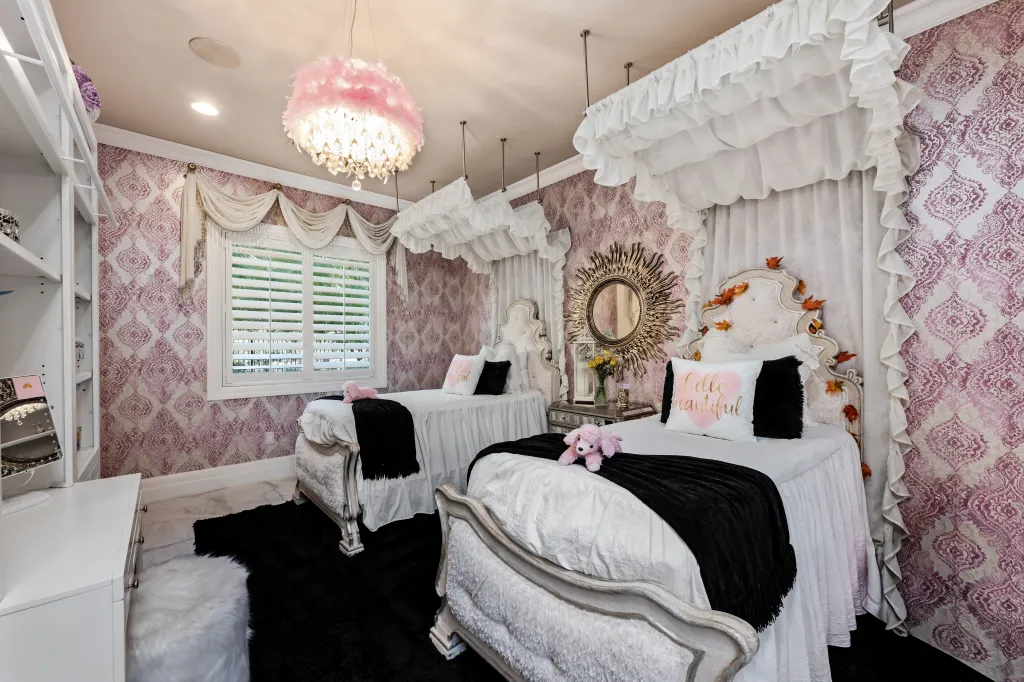 Bedroom with two beds, purple patterned wallpaper, a pink feathered chandelier, and a sunburst mirror.