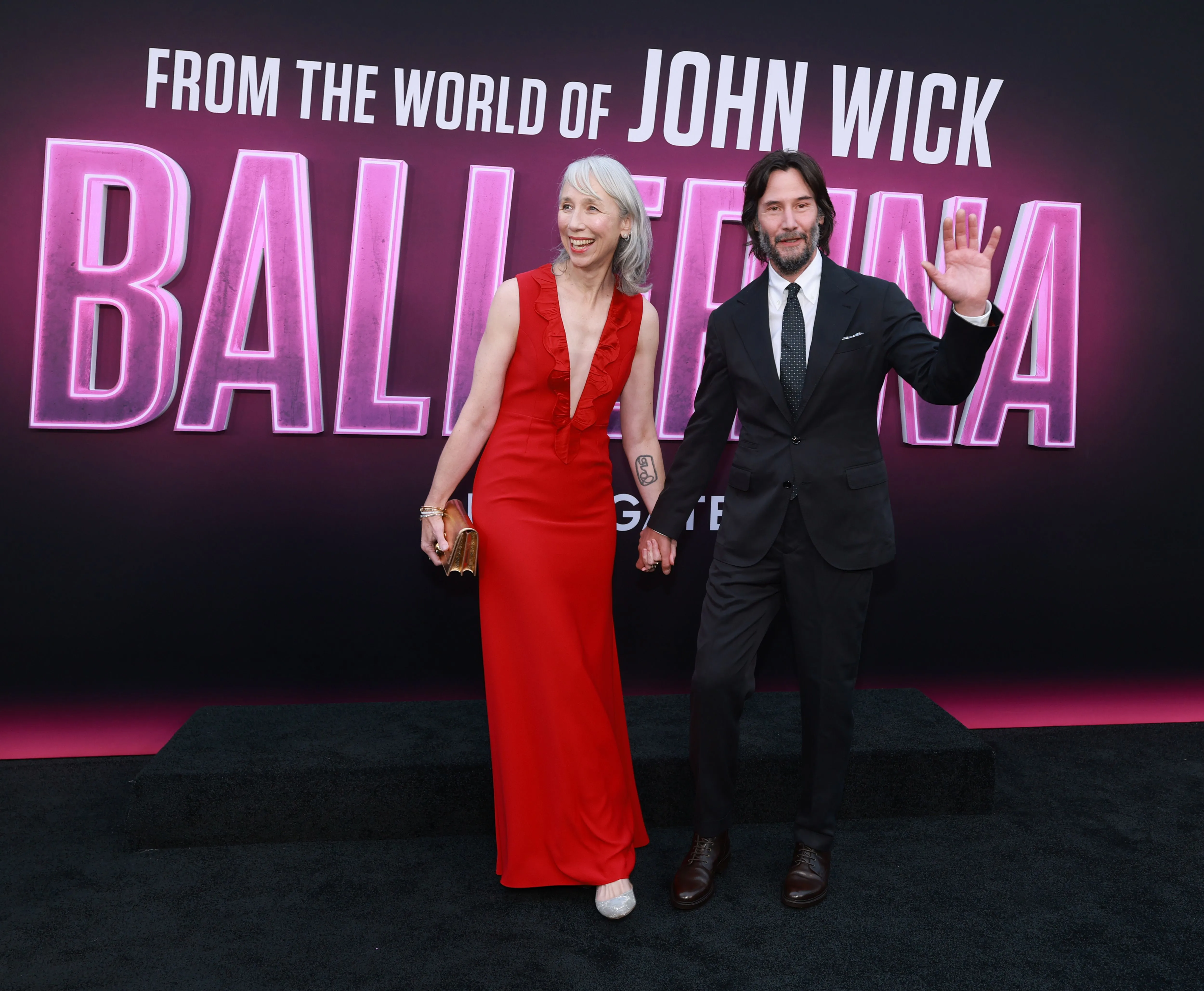 Alexandra Grant in a red dress and Keanu Reeves in a black suit holding hands at the 