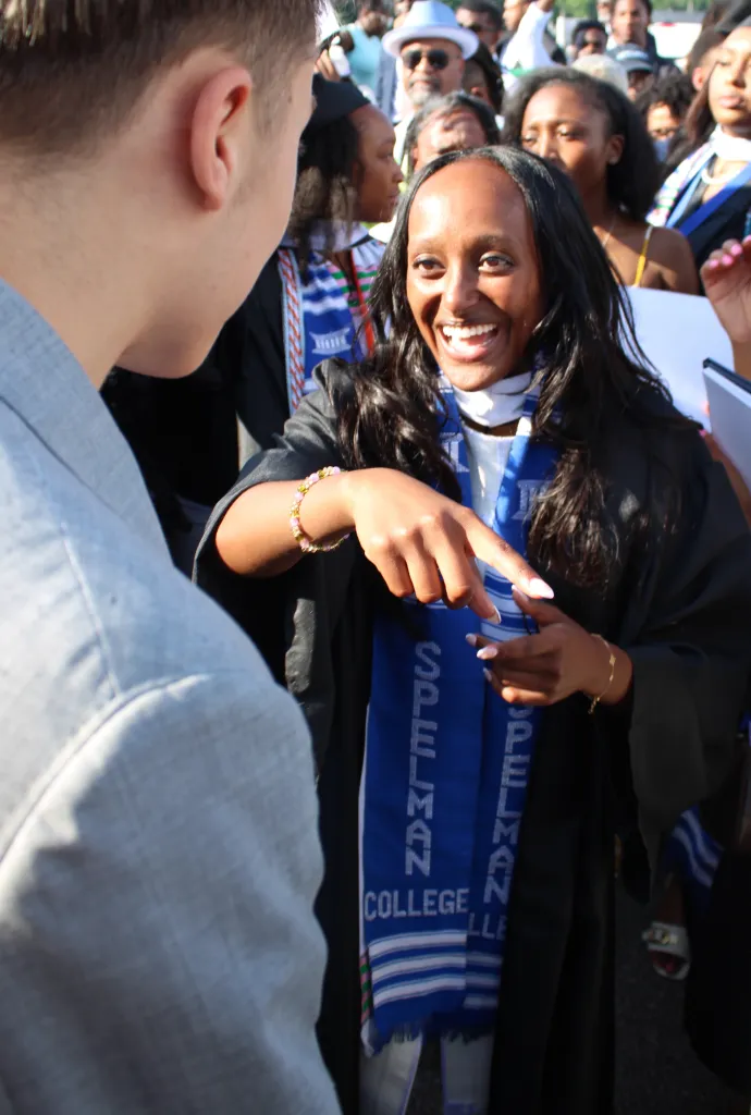 Zahara Jolie-Pitt in a graduation gown, wearing a blue and white Spelman College stole, pointing at someone in a light gray shirt.