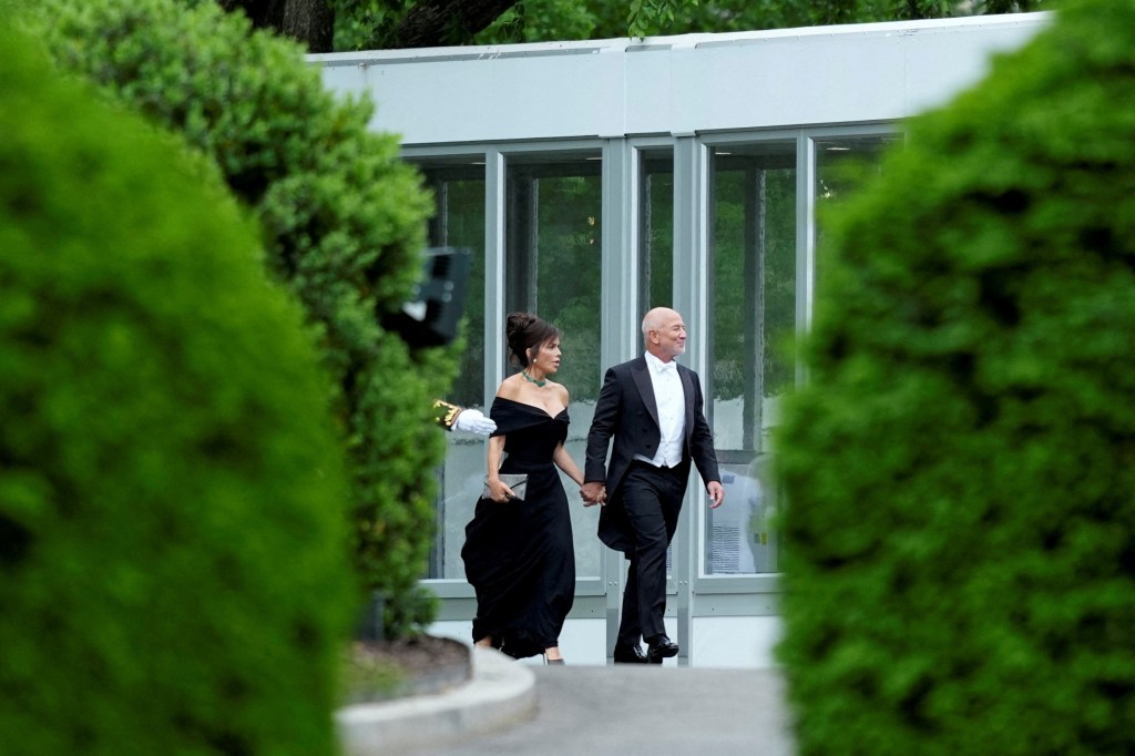 Jeff Bezos, in white tie, and Lauren Sanchez, in a black off-the-shoudler gown holding hands.