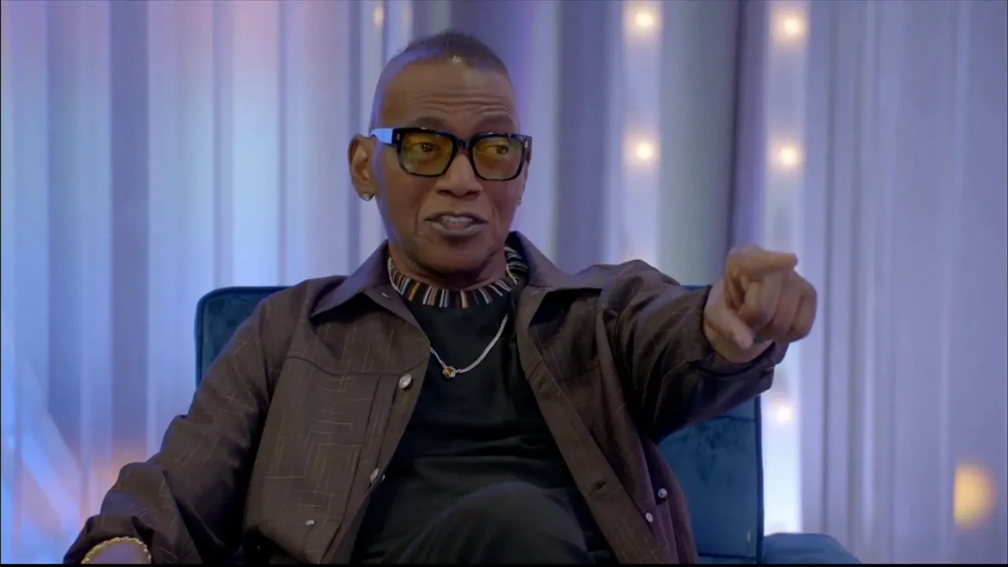 Randy Jackson sitting and pointing forward while wearing glasses and a striped shirt.