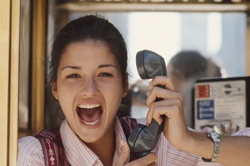 Janice Dickinson in a phone booth, holding a phone receiver and shouting with excitement.