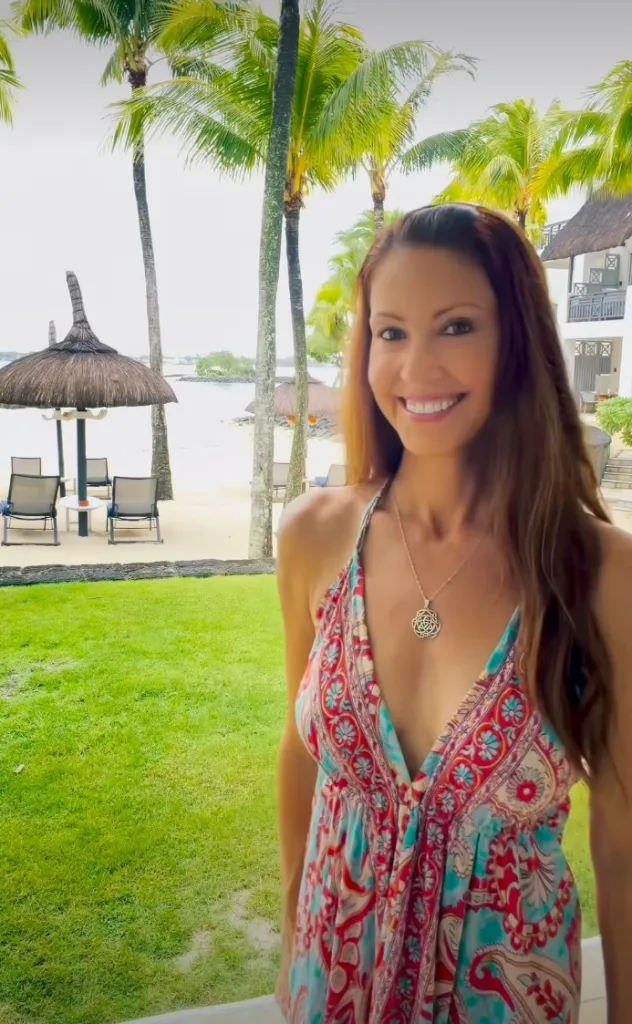 American Pie actress Shannon Elizabeth smiles at the camera, wearing a patterned dress, with palm trees and a beach in the background.