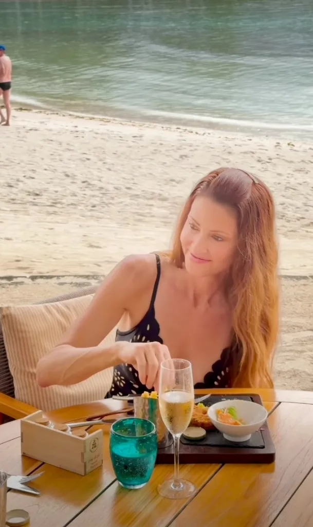 Shannon Elizabeth in a black bathing suit, seated at a table on a beach, with food and champagne in front of her.