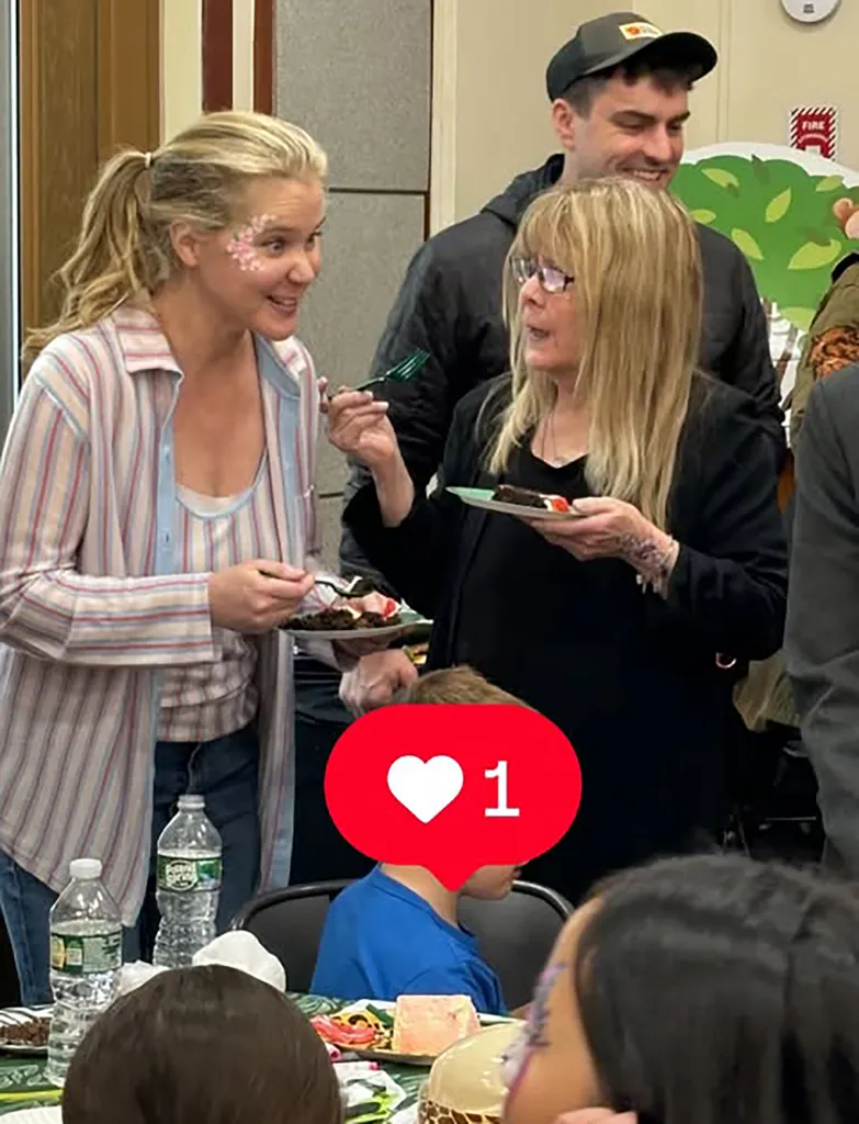 Amy Schumer smiling at another woman while holding a plate of dessert, with a man smiling in the background.