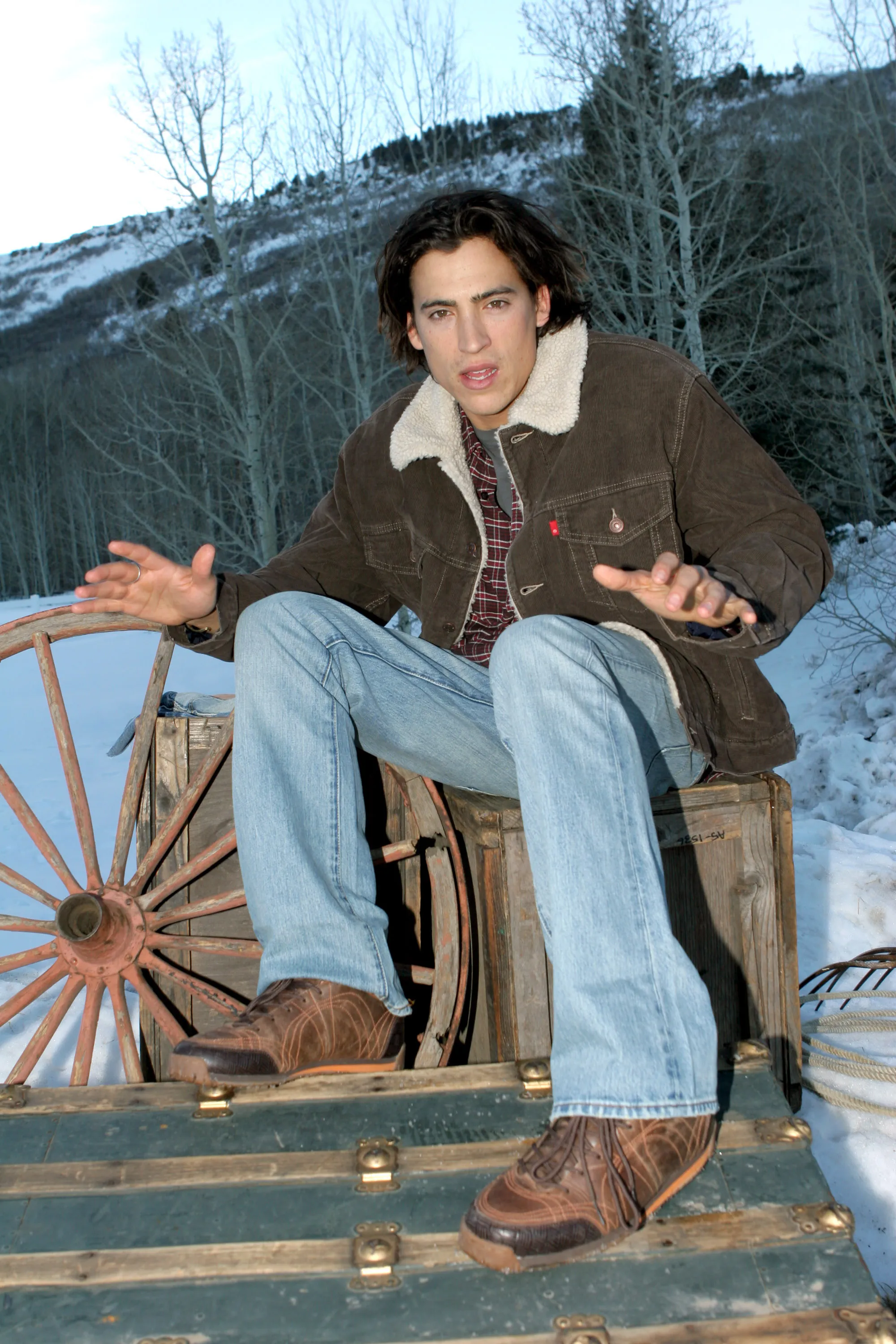 Andrew Keegan seated on a wooden crate in a snowy, outdoor setting with mountains and trees in the background.
