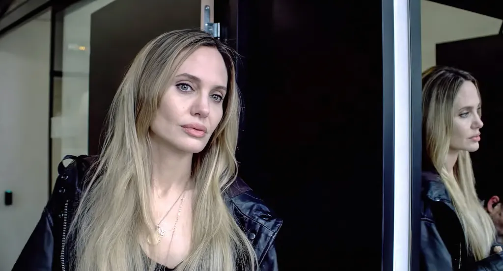 Portrait of Angelina Jolie with long blonde hair, wearing a black leather jacket, looking towards the viewer, with her reflection visible in a mirror on the right.