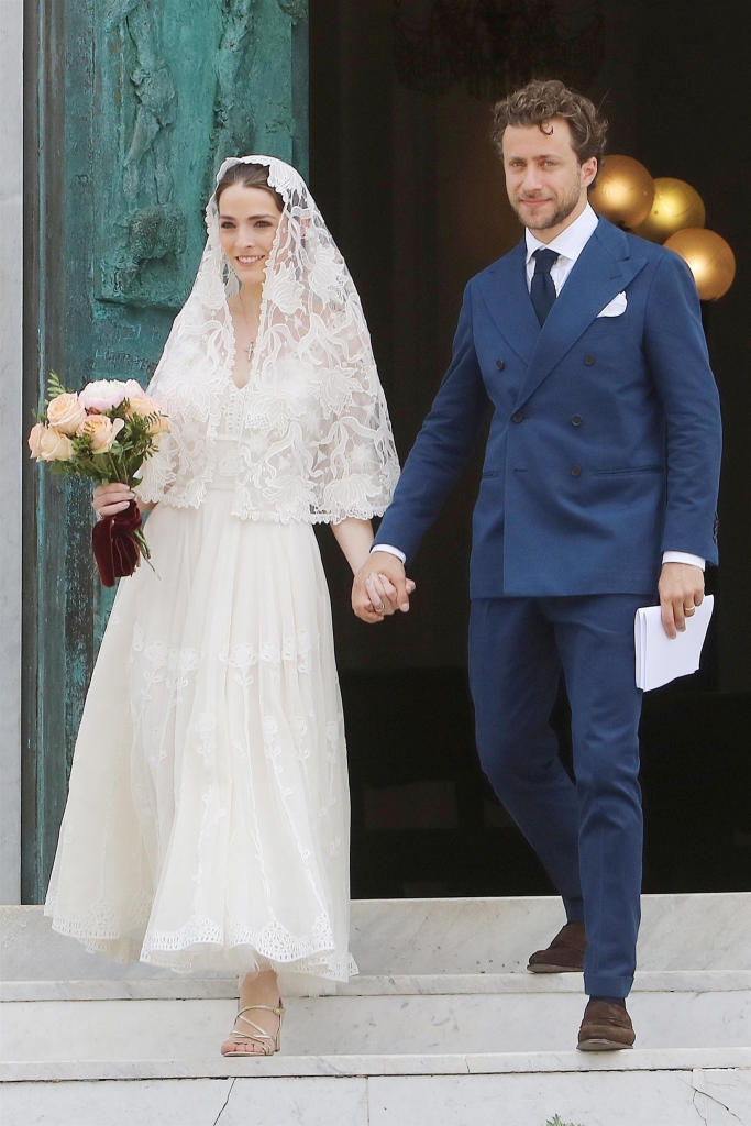 Francesco Carrozzini, in a blue suit and black tie and white shirt, and Bee Shaffer, in a white wedding gown and lace veil, and holding hands and smiling, descending steps in their wedding attire.
