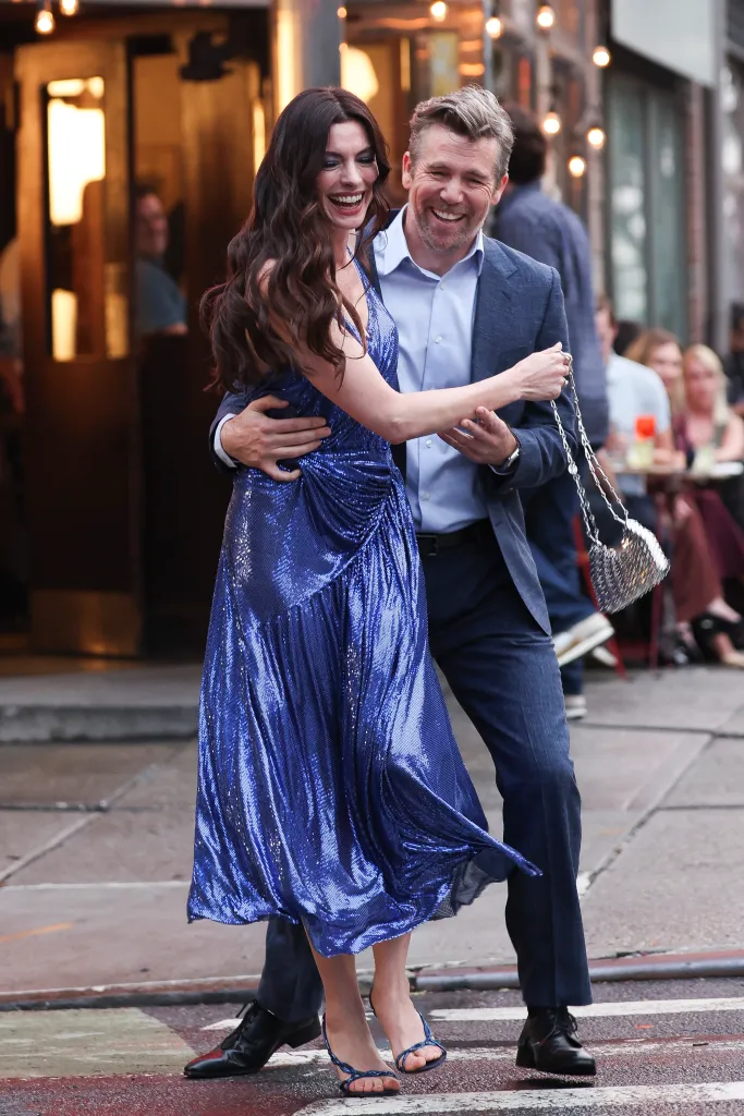 Anne Hathaway in a shimmery blue dress embracing Patrick Brammall, both laughing, on the set of 