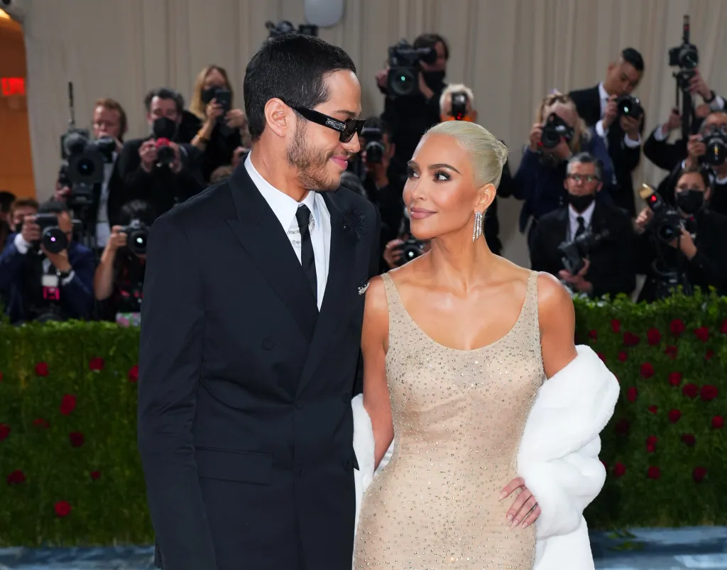 Pete Davidson in a black suit with sunglasses, and Kim Kardashian in a sparkling beige dress with a white fur wrap, look at each other at the Met Gala.