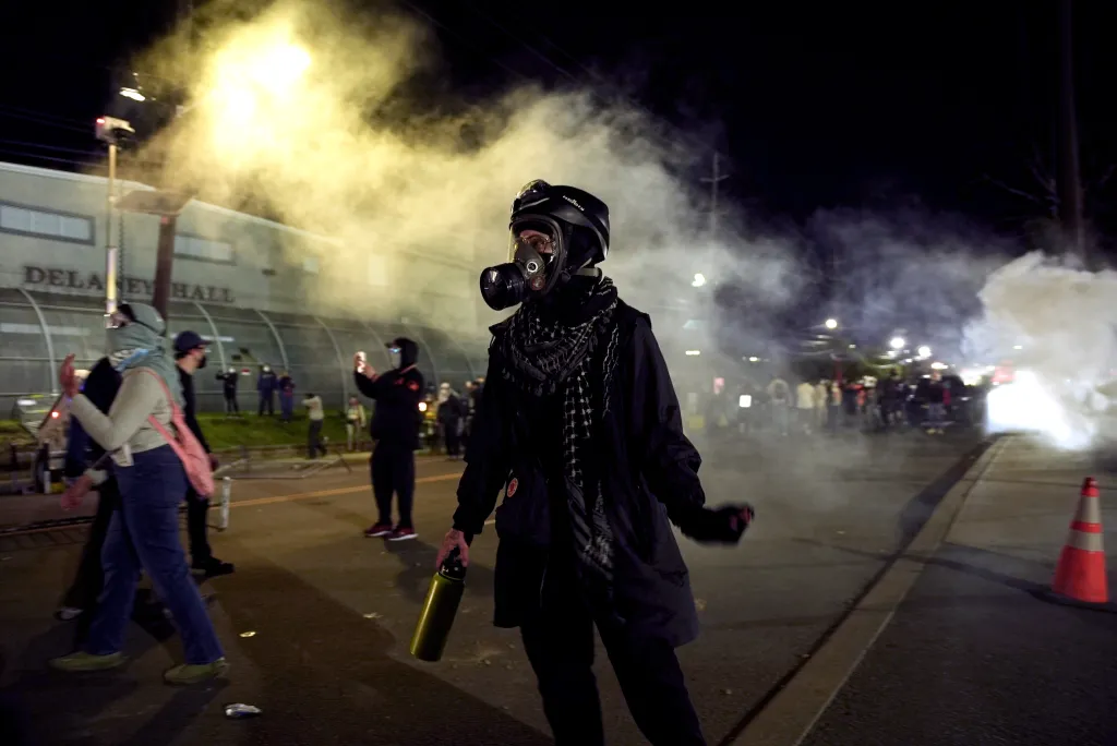 An anti-ICE protester in a gas mask and helmet holds a water bottle amid tear gas and other demonstrators.