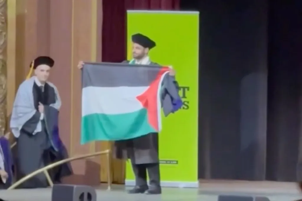 A graduate in cap and gown holds up a Palestinian flag on stage.
