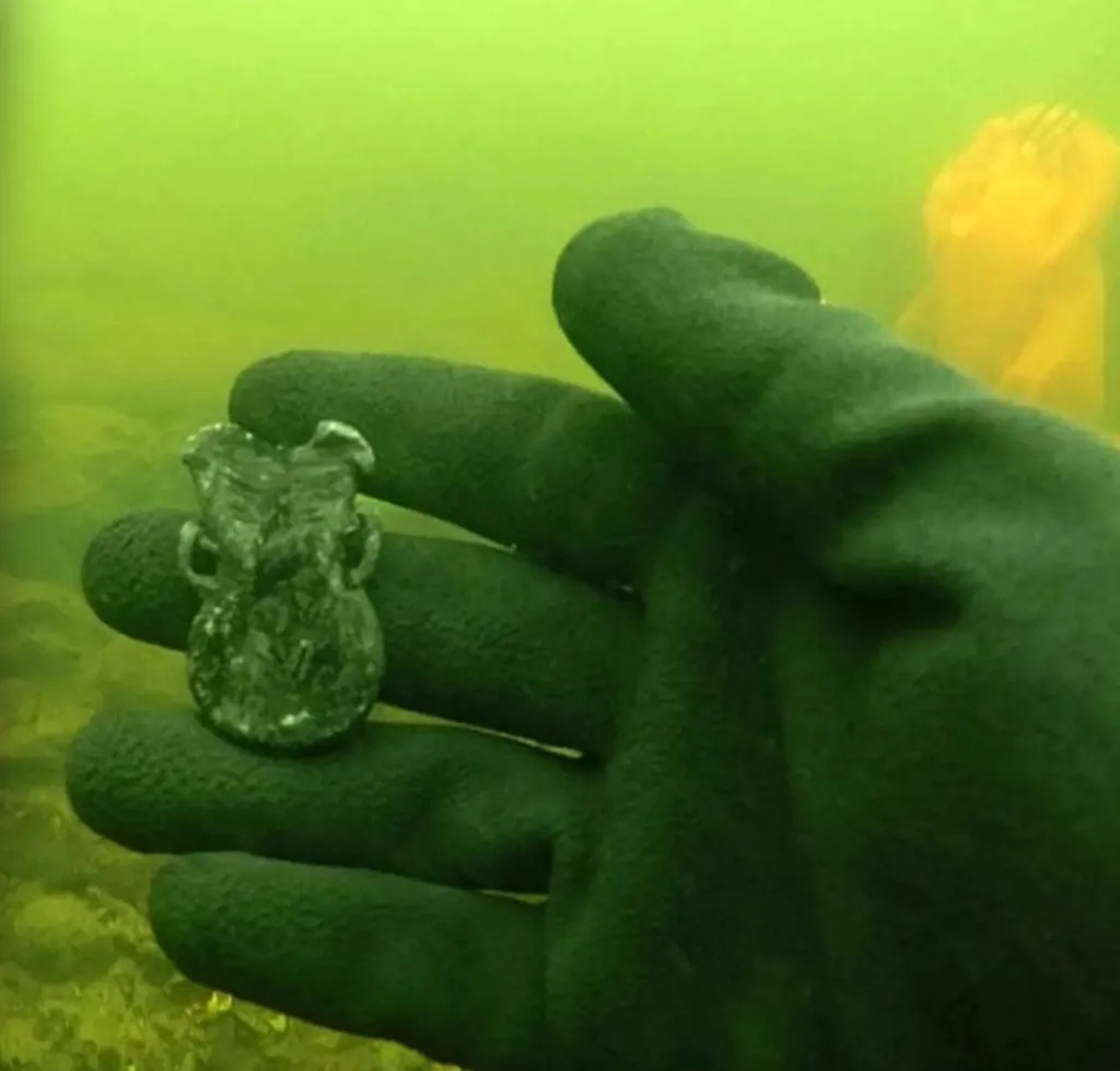 A gloved hand underwater holds a small, corroded religious artifact.