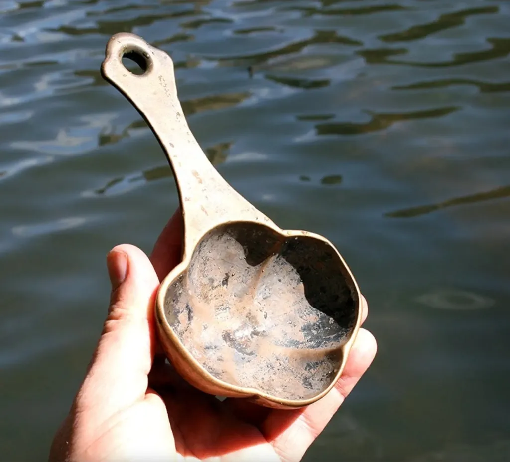A hand holds a silver christening spoon above water.