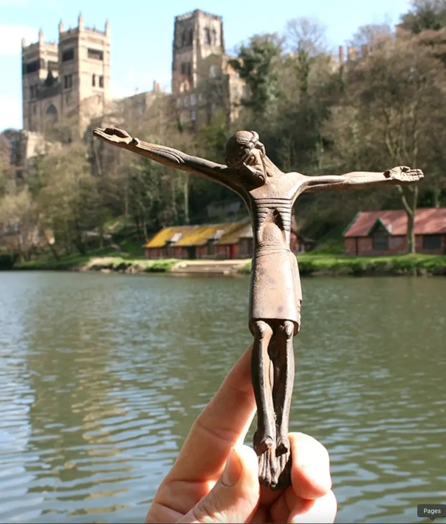 A hand holding a bronze crucifix in front of a river and a church.