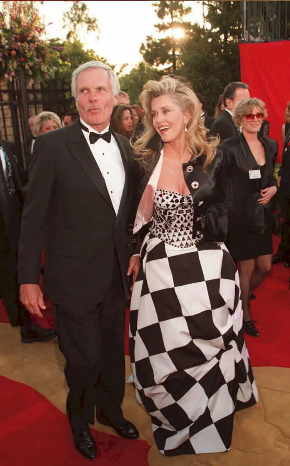 Jane Fonda, in a black and white checkered skirt and black jacket, poses with Ted Turner, in a black tuxedo, on a red carpet.