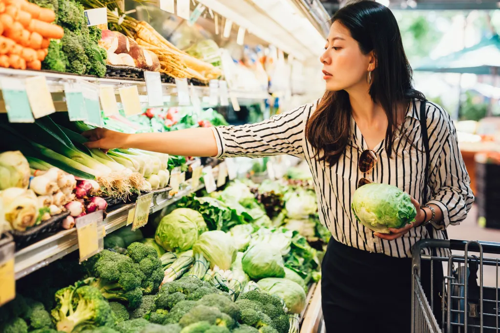 A woman in a striped shirt and black skirt shops for fresh produce in a supermarket, holding a head of lettuce and reaching for green onions.
