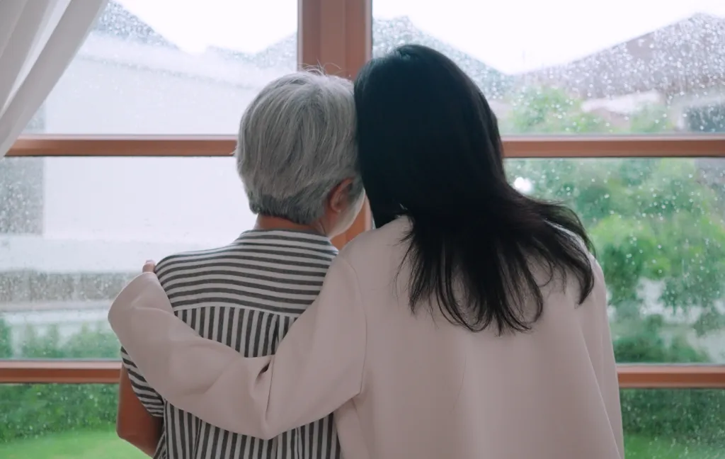 A daughter hugging her mother as they look out a window on a rainy day.