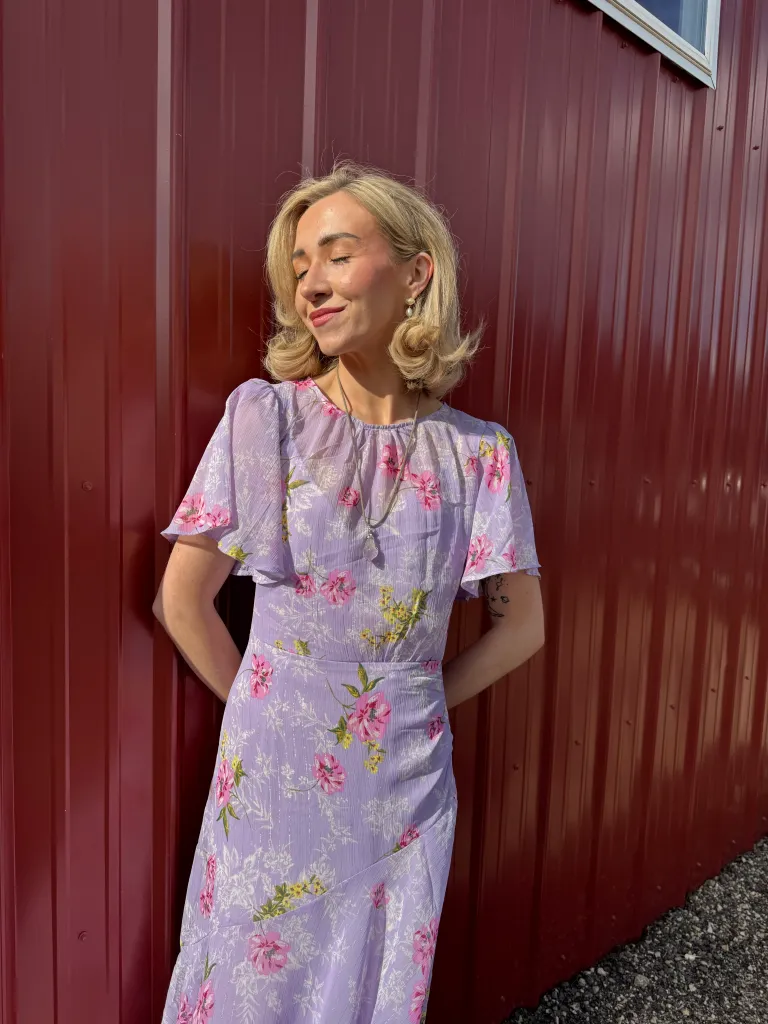 Jenna Lee Jane in a floral purple dress with pink flowers, smiling with her eyes closed against a red wall, in sunshine.
