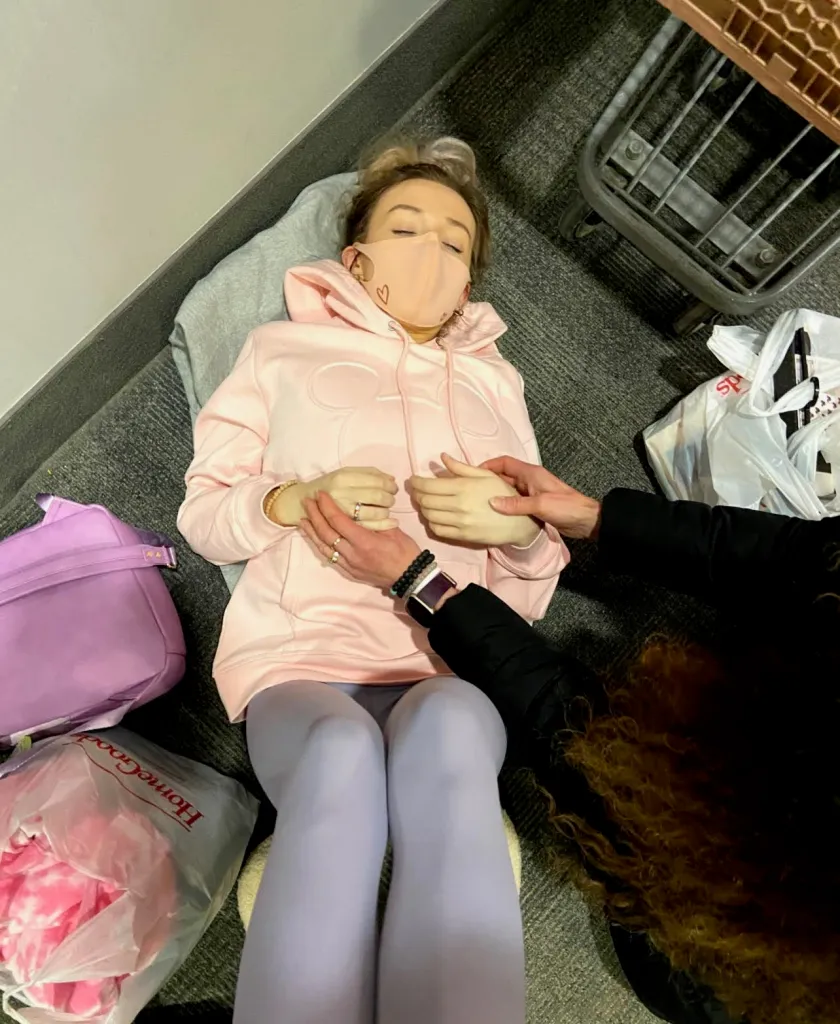 Jenna Lee Jane in a pink hoodie and face mask lies on the floor while another person holds their hands.
