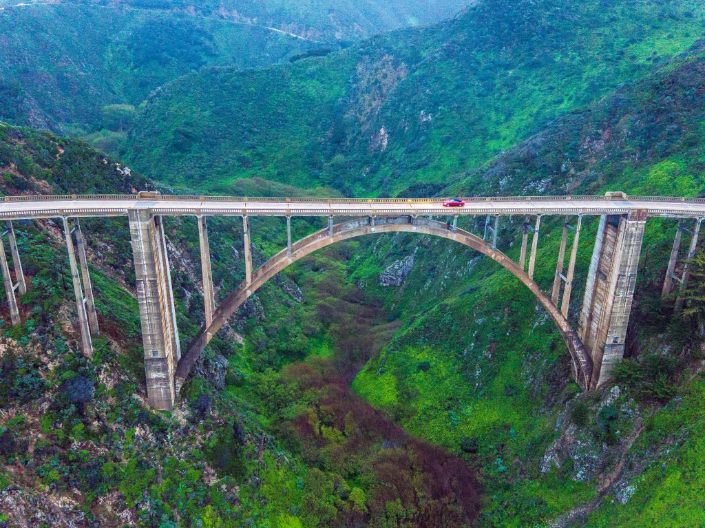 A red car drives over Bixby Bridge, which spans a deep canyon filled with green trees and bushes.