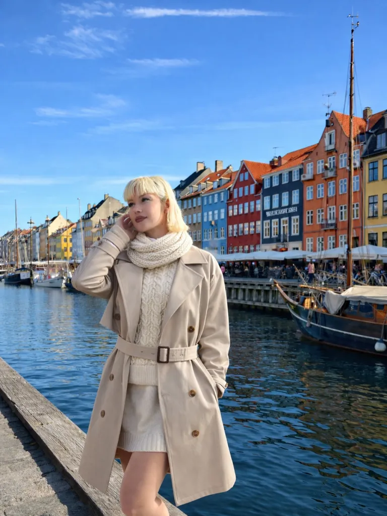 A woman in a beige trench coat and white knitwear poses by a canal with colorful buildings in the background.