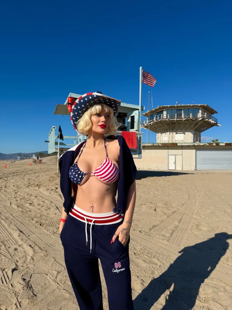 Aysegul Eraslan in a stars and stripes bikini top and hat, with navy pants, on a sandy beach.