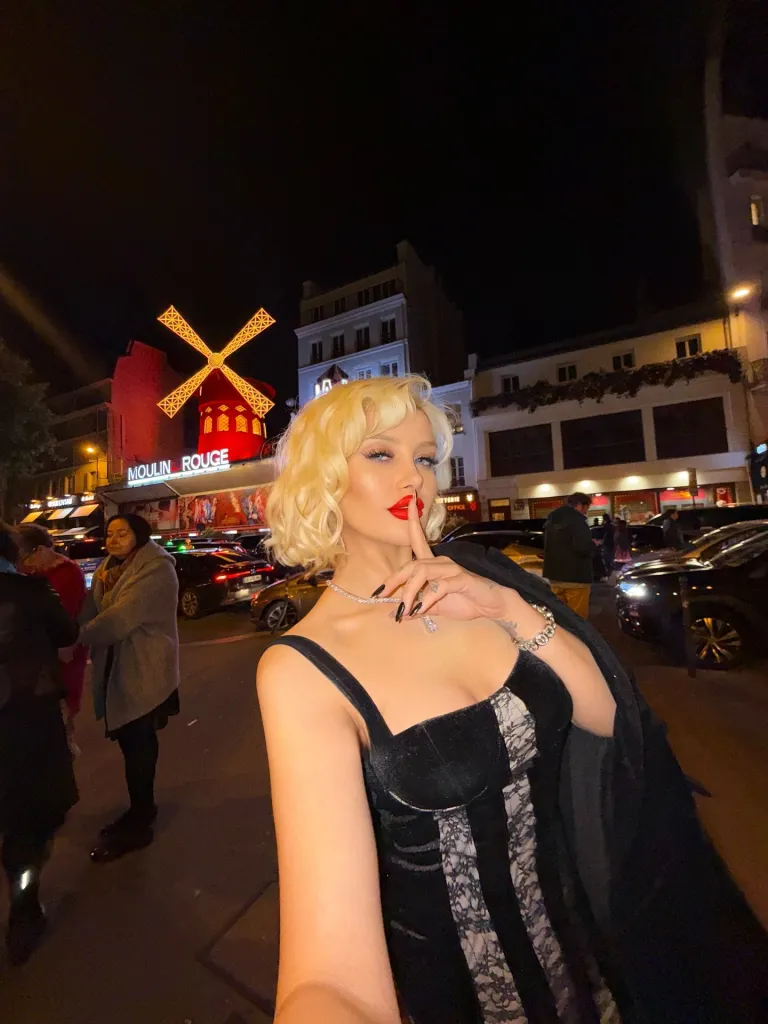 A woman in a black and white dress with blonde hair poses in front of the Moulin Rouge at night.