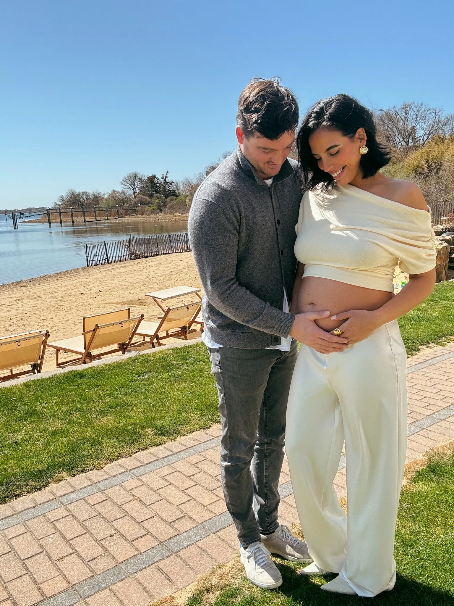 Man cradling a pregnant woman's belly with a beach and water in the background.